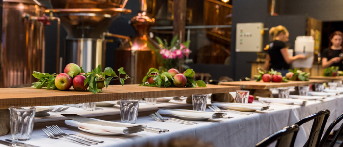Willie Smith’s Apple Shed 2 - credit Fred & Hannah Long dining table set with white tablecloths, glassware, and neatly arranged cutlery, featuring wooden centerpieces decorated with fresh apples and green foliage, in a room with large copper distilling equipment in the background.