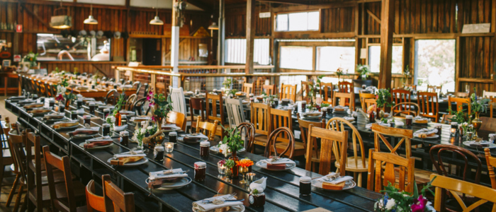 Willie Smith’s Apple Shed 3 - credit Jonathan Wherrett Rustic indoor dining space with long wooden tables set for a meal, featuring plates, cutlery, glassware, and small floral centerpieces, surrounded by a mix of wooden chairs in a warm, timber-paneled room with large windows.
