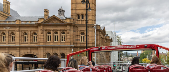 Passengers seated on a red double-decker tour bus observe the scenery. In the background, a sandstone clock tower and ornate historic building dominate the view, with traffic and city streets stretching into the distance.