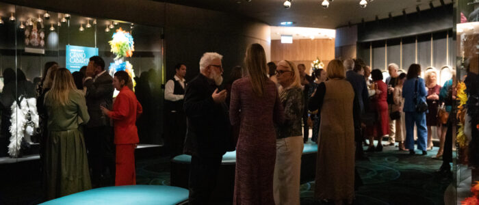 A lively indoor event space with guests mingling and chatting. Attendees are dressed in elegant outfits, while the room is decorated with illuminated display cases showcasing costumes and modern ceiling lights. A teal bench is in the foreground.