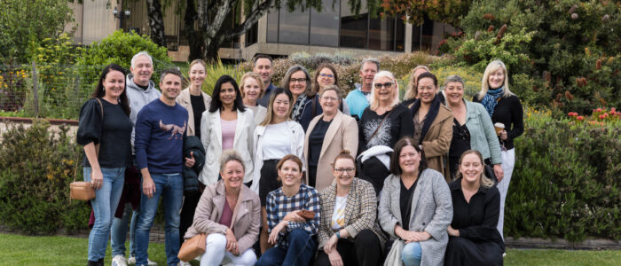 A large group of people gathered for a photo outdoors, standing and crouching on grass. Behind them, there are trees, shrubs, and a modern building with reflective windows. Most of the group members are smiling and dressed in relaxed clothing.