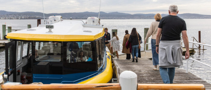 A yellow and white boat is docked at a wooden pier while a group of people, including a man in a black shirt and gray sweater, walk toward the end of the pier. The water and distant hills form a calm backdrop under an overcast sky.