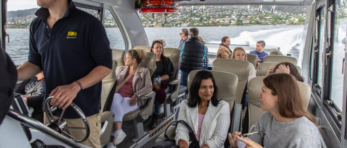 Passengers sit inside a modern boat cabin, engaging in conversation and looking out at the water. A man in a navy shirt steers the boat, while the wake behind shows movement. Large windows reveal a hilly shoreline with houses in the distance.