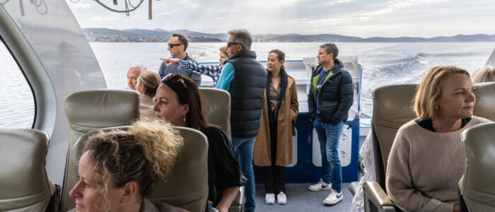 Passengers inside a boat cabin, some seated while others stand near the back observing the view. Sunlight streams through the windows, illuminating the calm water and distant hills outside.
