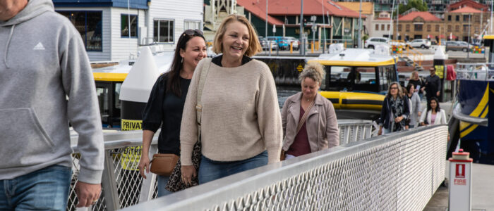Several people walking down a pier toward the camera, led by a cheerful woman wearing a beige sweater. Behind her, two other women walk closely together, with additional individuals further back. The harbor setting features a docked yellow and white boat, waterfront buildings, and a cloudy sky.