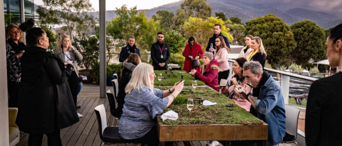 A group of people gather on a modern outdoor terrace with wooden floors and sleek architectural features. Guests sit and stand around a moss-covered table, enjoying drinks and conversations, while mountains and greenery form a scenic backdrop.