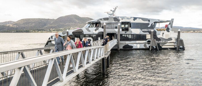 Passengers exit or board a sleek, camouflaged ferry named "MONA ROMA" via a long, elevated walkway over the water. The calm water and mountains in the background are visible under a cloudy sky.