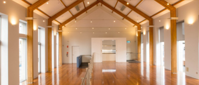 The Waterside Pavilion at Mawson Place 1 A bright and spacious interior of a hall with polished wooden floors, exposed beams, and large windows letting in natural light.