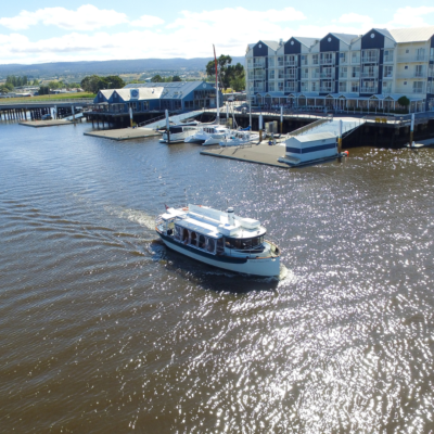 A scenic view of the Tamar River featuring a boat with an open cabin sailing past a marina. The docked yachts and waterfront buildings, including apartments and a boathouse, line the background.
