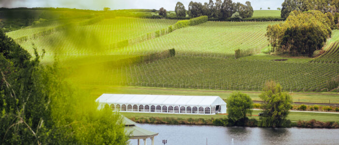 A very large white tent marquee in the distance next to a lake with a gazebo and swans. Behind the marquee is rolling green fields of vineyard and trees