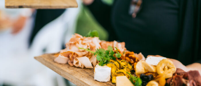A close-up of a charcuterie board being held by a person dressed in black. The board includes slices of cured meats, cheeses, fresh herbs, and small servings of accompaniments like dried fruits and nuts.