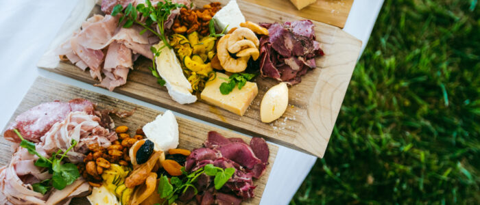 Three wooden charcuterie boards arranged on a white table, topped with a variety of meats, cheeses, roasted nuts, dried fruits, and fresh greens. The boards are set outdoors on a lawn.