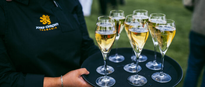 A server wearing a black apron embroidered with "Josef Chromy Tasmania" holds a tray of sparkling wine glasses outdoors. Each glass is filled with golden bubbly, with a green lawn visible in the background.