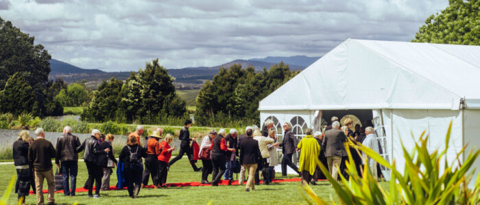 Guests mingling outdoors at an event, standing on a red carpet leading to a white marquee tent, surrounded by lush greenery and distant hills.