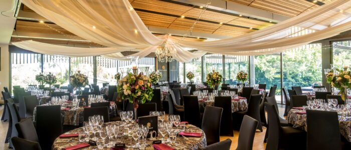 An elegant banquet hall with round tables covered in patterned tablecloths, decorated with floral centerpieces and surrounded by black chairs, under draped fabric and chandeliers.