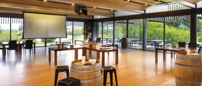 A bright, spacious room with wooden floors, a large projector screen, and barrel-style tables, surrounded by large windows overlooking greenery.