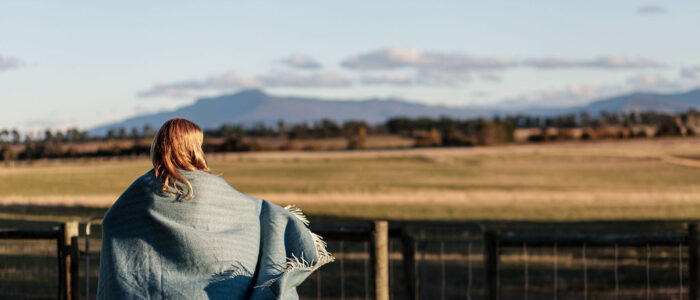 Woman looking out over a field of grass with a blue blanket over her shoulders covering her entire back.