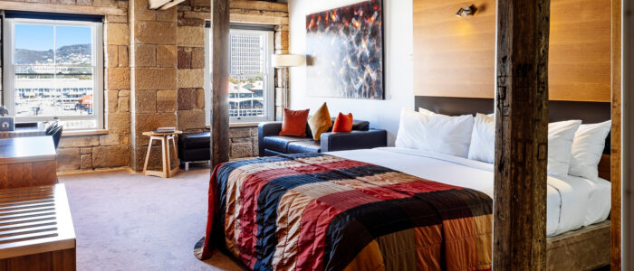 A rustic-modern hotel room with a king-size bed, a patchwork quilt, and a cozy seating area. Large windows framed by stone walls provide natural light and a view of a harbor and hillside buildings. A contemporary painting hangs above the couch, adding an artistic touch.