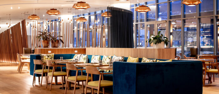 Interior of a restaurant. Wooden floorboards with a blue couch that surrounds small circular tables set with salt and pepper shakers. Roof is a plain white with bronze hanging lights. The far wall is made of large windows overlooking the city