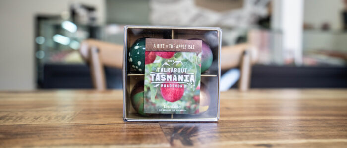 A small box with four handcrafted chocolates sits on a wooden countertop. On the box is packaging that says "Talk About Tasmania. Roadshow"