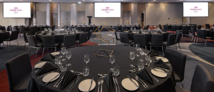 A large event space set up for an event. Scattered around the room are large circular tables set on black tablecloths, with a golden cube-wire ornament in the center. On the back wall are three projector screens with the words "Crowne Plaza, an IHG hotel Hobart" displayed
