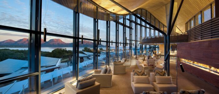 A spacious lounge with modern beige armchairs and a sleek fireplace, illuminated by warm lighting. Floor-to-ceiling glass windows offer a stunning view of distant mountains and a serene body of water at sunset.