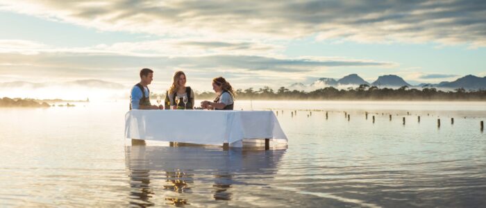 A unique dining experience unfolds as three friends sit at a white-tableclothed table set in ankle-deep water, sipping wine with mist-covered mountains in the background.