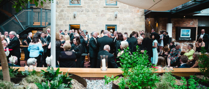 lively gathering of well-dressed guests in an open-air courtyard with exposed stone walls and lush greenery. Attendees in black-tie attire mingle, chat, and enjoy drinks under a stretched fabric canopy, while a staircase and a balcony add architectural interest.