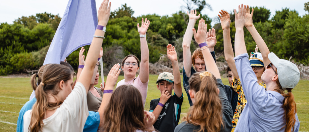 A group of young people stand in a circle on a grassy field, raising their hands in unison as part of a group activity. Most are wearing purple wristbands, and one person holds a light purple flag. Trees and shrubs form a natural background, suggesting an outdoor camp or team-building event.