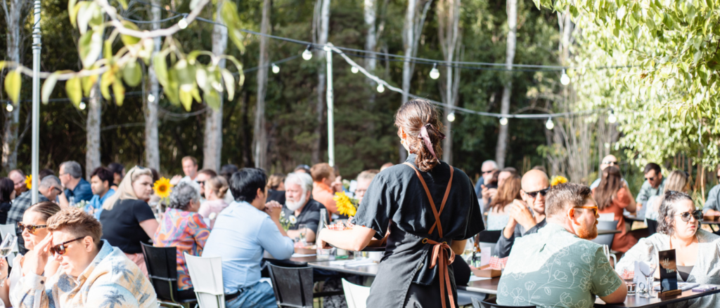 A busy outdoor dining event takes place in a forested setting, with guests seated at long tables under string lights. A server in a black apron carries a tray through the crowd. Sunflowers decorate the tables, and sunlight filters through the trees, creating a warm and festive atmosphere.