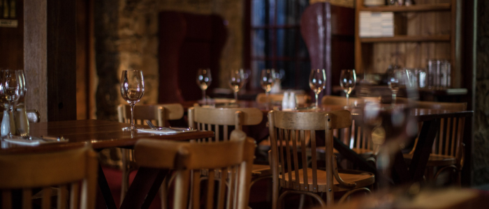 The interior of a restaurant or dining area with wooden tables and chairs. The tables are set with wine glasses, cutlery, and napkins. The background includes wooden shelves and a large armchair. The lighting is warm, creating a cozy atmosphere.