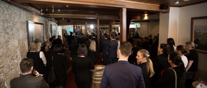 A group of people gathered in a room, possibly for an event or meeting. The room has stone walls on one side and wooden elements, including the ceiling and pillars. There are paintings on the walls and a screen at the front where some people appear to be giving a presentation or speech. The attendees are standing and facing towards the front of the room.