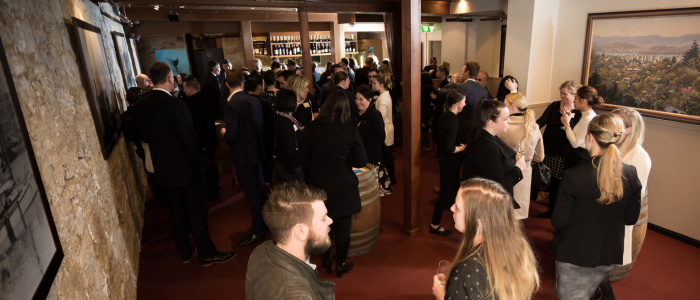 A group of people gathered in what appears to be a social or networking event. The setting is indoors, with wooden beams and wine bottles visible on shelves in the background. There are framed pictures on the walls, and the attendees are engaged in conversation, standing in small groups throughout the room.