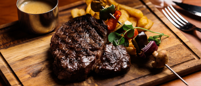 A wooden serving board with a cooked steak, a skewer of vegetables (including mushrooms, red onion, and bell peppers), some fried potato wedges, and a small metal cup containing sauce. Utensils (a fork and knife) are placed on the right side of the board.
