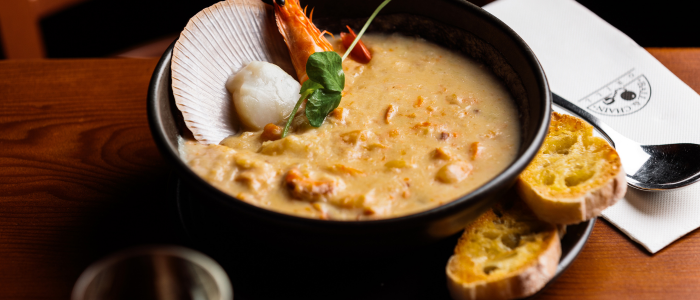 A bowl of creamy seafood soup garnished with a large shrimp, a scallop, and a green herb. The bowl is placed on a wooden table next to two slices of toasted bread. There is also a white napkin with some text and an emblem partially visible, along with a spoon resting on the napkin.