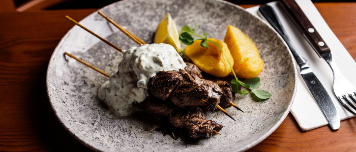 A plate of food with skewered grilled meat topped with a creamy sauce, accompanied by two potato wedges and a lemon wedge. There is also some green garnish on the plate. The plate is placed on a wooden table, and next to it are a fork and knife on top of a white napkin.