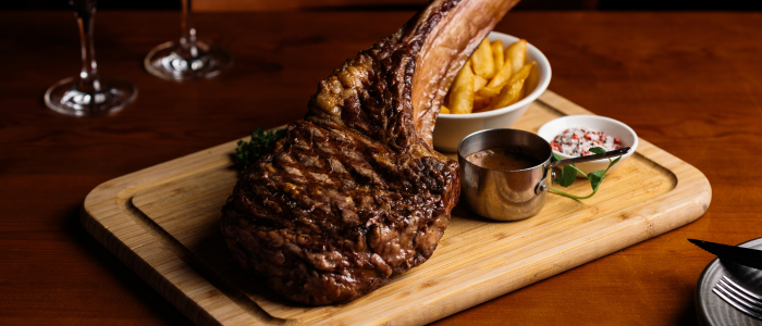 A large, grilled tomahawk steak served on a wooden cutting board. Accompanying the steak are a bowl of French fries, a small metal cup of sauce, and another small dish containing what appears to be a garnish or seasoning. The setting includes two wine glasses in the background and part of a knife visible on the right side of the image.