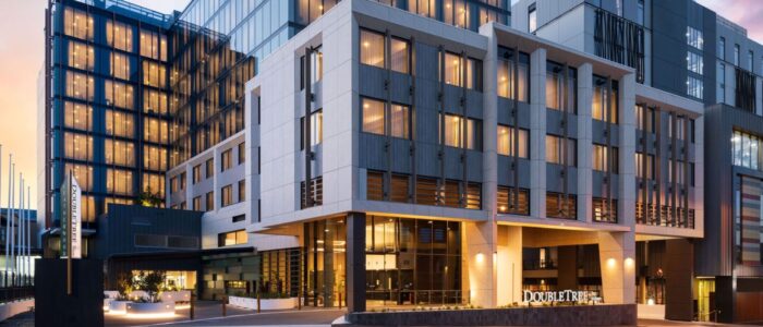 The DoubleTree by Hilton Hobart exterior. It's a modern, multi-story building complex with large glass windows and contemporary architectural design. The exterior is well-lit, suggesting it is either early morning or evening.