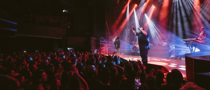 A live music performance on stage with a large audience. There are multiple performers, including a singer and a keyboard player, under colorful stage lighting. The audience is engaged, with many people raising their hands and taking photos or videos.