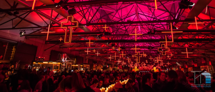 An indoor venue with a high ceiling and exposed beams, illuminated by numerous red neon cross-shaped lights hanging from the ceiling. The space is crowded with people, and there are various booths or stalls along the sides. The overall ambiance is vibrant and lively, with a focus on the striking red lighting.