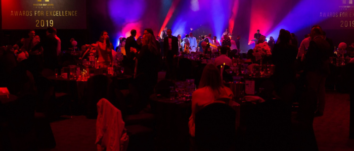 A dimly lit event with people gathered around tables, likely attending an awards ceremony. There are colorful lights in the background, and a stage with performers or speakers. The text on the left side of the image reads 'AWARDS FOR EXCELLENCE 2019.'