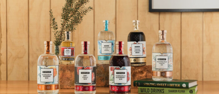 A collection of various bottles of spirits arranged on a wooden surface. There are eight bottles in total, each with distinct labels indicating different types of gin, vodka, and other spirits. Behind the bottles, there is a sprig of greenery placed in one of the bottles. To the right side of the image, there are two books stacked on top of each other titled 'Strong, Sweet & Bitter' and 'Wild Drinks' by Sharon Flynn. The background consists of wooden paneling and part of a framed picture is visible on the wall.