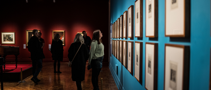 A group of people in an art gallery with framed pictures displayed on the walls. One wall is painted blue and another red. Several individuals are closely observing the artwork.