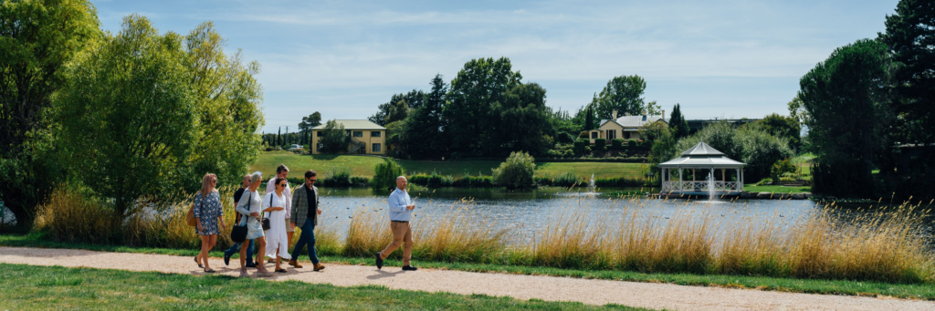 A group of people walking along a path next to the lake Josef Chromy. The background features lush greenery, trees, and several buildings. There is also a white gazebo near the water's edge on the right side of the image. The sky is clear with some clouds, indicating pleasant weather.