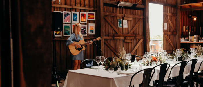 A rustic-style room with wooden walls, ceiling beams and hanging lights. A woman in a blue dress stands playing the guitar to the left of the image close to a wall with ten small paintings. In the middle of the room is a long table with a white table cloth, wine glasses and native Australian plants assorted into a decorative arrangement.