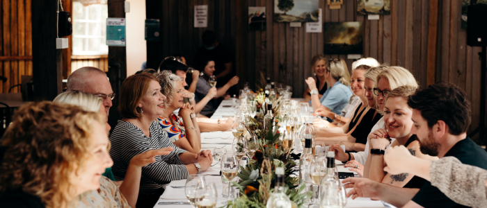 A long dining table with people seated on both sides, engaged in conversation. The table is set with wine glasses, plates, and cutlery. There is a floral centerpiece running along the middle of the table. The background features wooden walls with various framed pictures hanging on them.