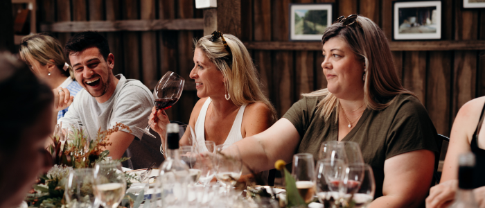 A group of people laughing and enjoying a social gathering at a long dining table. A blonde woman in a white top sits in the foreground, mid-laugh, while others around her are also smiling. The rustic wooden walls are adorned with framed artwork.