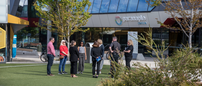 A group of people standing outside a building with the sign 'paranaple centre.' The building features large glass windows, and the surrounding area includes trees and greenery.