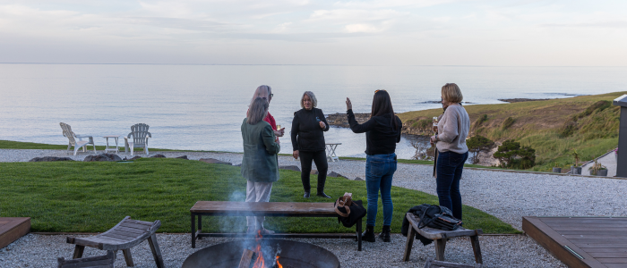 Four people standing near a fire pit with benches around it, facing the ocean. The outdoor setting includes a grassy area, a gravel path, and some chairs in the background. The sky is partly cloudy, and the ocean appears calm.