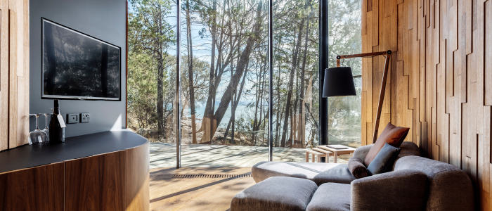 A modern, cozy living room with floor-to-ceiling windows overlooking a forested area. The room features wooden walls and flooring, a gray armchair with an ottoman, a black floor lamp, and a flat-screen TV mounted above a wooden console. Natural light fills the space, creating a warm and inviting atmosphere.